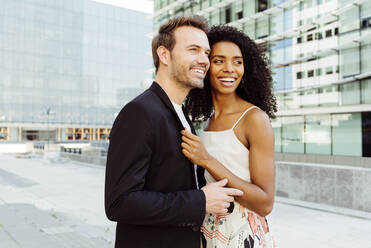 Seitenansicht einer attraktiven schwarzen Frau, die lächelt und einen kaukasischen Mann umarmt, während sie gemeinsam auf der Straße stehen - ADSF03289