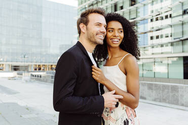 Side view of attractive black woman smiling and embracing Caucasian man while standing on city street together - ADSF03289