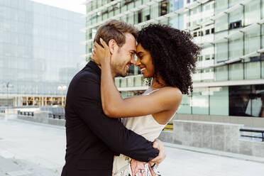 Side view of attractive black woman smiling and embracing Caucasian man while standing on city street together - ADSF03290