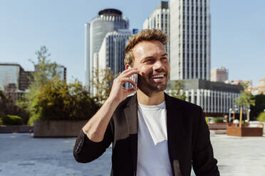 Attraktiver junger Mann, der lächelnd sein Smartphone benutzt, während er auf der Straße einer modernen Stadt an einem sonnigen Tag steht - ADSF03295
