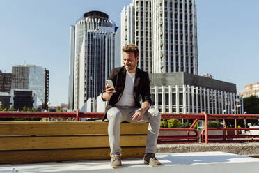 Handsome guy smiling and browsing smartphone while sitting on bench on street of modern city - ADSF03296