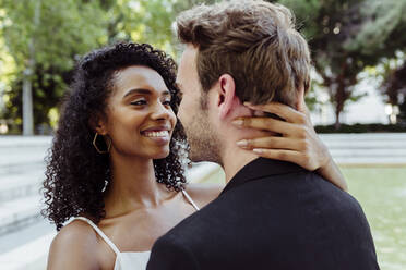 Attractive African-American woman smiling and looking at Caucasian man while standing in park and touching his neck - ADSF03312