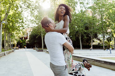 Side view of handsome Caucasian man lifting laughing African-American woman while standing on park alley on sunny day - ADSF03317