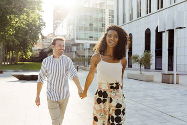 Hübsche schwarze Frau und kaukasischer Mann lächelnd und die Hände haltend beim Gehen auf der Stadtstraße zusammen an einem sonnigen Tag - ADSF03320