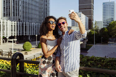 Beautiful black woman and handsome Caucasian man posing for selfie while standing on background of modern city - ADSF03321