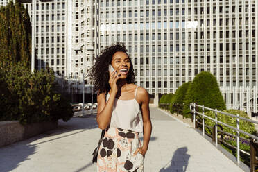 Attraktive afroamerikanische Frau, die lacht und mit ihrem Smartphone spricht, während sie an einem sonnigen Tag durch die Stadt läuft - ADSF03325