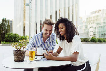 Nette multirassische Paar lächelnd und Surfen modernen Smartphone beim Sitzen am Tisch in schönen Outdoor-Café zusammen - ADSF03327