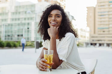 Charmante afroamerikanische Frau, die lächelt und wegschaut, während sie ein Glas mit einem leckeren Getränk in der Hand hält und sich auf einen Tisch in einem Straßencafé stützt - ADSF03332