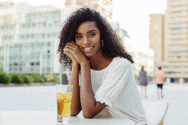 Charmante afroamerikanische Frau, die lächelt und wegschaut, während sie ein Glas mit einem leckeren Getränk in der Hand hält und sich auf einen Tisch in einem Straßencafé stützt - ADSF03333