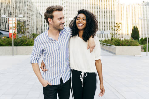 Cheerful black woman and Caucasian man hugging and laughing while walking on city street together - ADSF03335