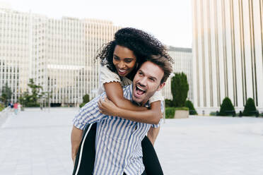 Handsome Caucasian man giving piggyback ride to African-American woman while having fun on city street together - ADSF03342