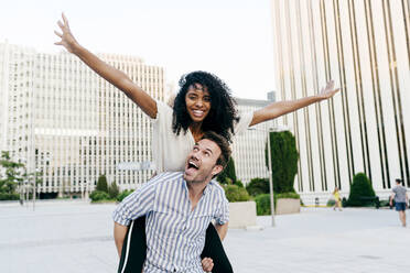 Handsome Caucasian man giving piggyback ride to African-American woman while having fun on city street together - ADSF03343