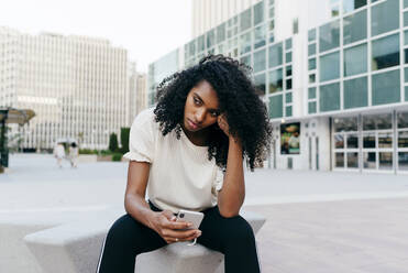 Junge afroamerikanische Frau reibt sich die Schläfe und benutzt ihr Smartphone, während sie auf der Straße einer modernen Stadt sitzt - ADSF03345