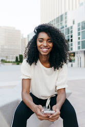 Hübsche afroamerikanische Frau lächelt und schaut in die Kamera, während sie auf der Straße sitzt und auf einem modernen Smartphone surft - ADSF03346