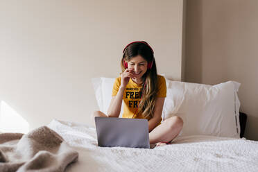 Fröhliche hübsche Frau mit Kopfhörern sitzt zu Hause auf dem Bett und benutzt einen Laptop. - ADSF03406