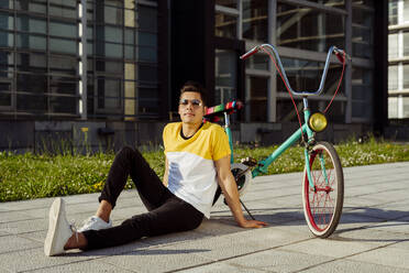 Attraktiver Mann mit Brille sitzt auf dem Boden in der Nähe von bunten Vintage-Fahrrad und schaut in die Kamera - ADSF03448