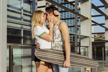 Attraktives junges Paar steht in der Stadt und hält Skateboard umarmt und küsst - ADSF03458