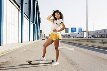 Lovely ethnic woman in nice sportswear stepping on skateboard and looking at camera while standing in middle of empty road in city - ADSF03482
