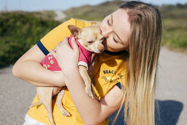 Glückliches lässiges Mädchen mit langem blondem Haar, das einen kleinen Hund (Chihuahua) hält und küsst, der auf der Straße im Sonnenschein steht. - ADSF03522