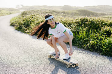 Cheerful casual girl in sunglasses with hair waving riding long board on paved road in nature. - ADSF03525