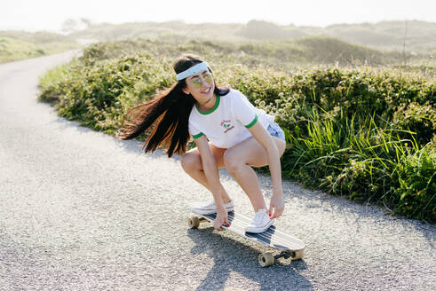 Fröhliches, lässiges Mädchen mit Sonnenbrille und wehenden Haaren auf einem Longboard auf einer asphaltierten Straße in der Natur. - ADSF03525