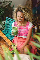 Schöne glückliche schwarze junge Frau hält ein Glas Shake und sitzt auf einem Stuhl in der Nähe von Tisch mit Schüssel Gemüse in Mexiko - ADSF03550