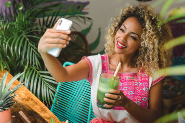 Schöne glückliche schwarze junge Frau mit Glas Shake nehmen selfie auf Handy und sitzen auf Stuhl in der Nähe von Tisch zwischen Pflanzen in Mexiko - ADSF03553