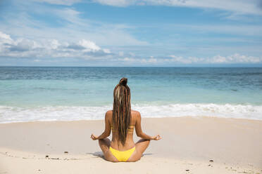 Rückenansicht einer attraktiven Frau im Bikini, die auf Sand am Meer in Bali, Indonesien, sitzt und meditiert - ADSF03565