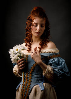 Hübsche Frau in mittelalterlichem Kleid hält die Augen geschlossen und hält einen Strauß weißer Blumen, während sie auf schwarzem Hintergrund steht - ADSF03567