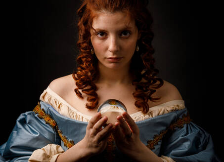 Schöne Frau in elegantem mittelalterlichem Kleid, die eine kleine Glaskugel hält und in die Kamera schaut, während sie auf schwarzem Hintergrund steht - ADSF03568
