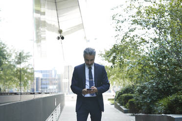 Geschäftsmann schreibt eine SMS über sein Smartphone, während er vor einem Büro in der Stadt steht - PMF01231