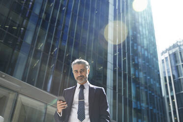 Lächelnder Unternehmer mit Mobiltelefon vor einem Bürogebäude in der Stadt an einem sonnigen Tag - PMF01252