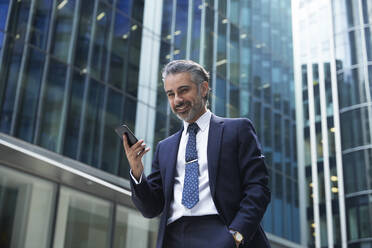 Smiling businessman using mobile phone outside office building in city - PMF01254