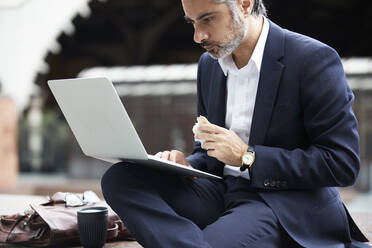 Businessman using laptop while eating food in city - PMF01275