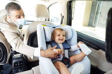 Vater trägt Schutzmaske Baby Junge sitzt in Kindersitz in einem Auto - JCMF01040