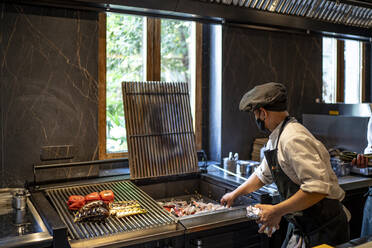 Koch mit Schutzmaske bei der Zubereitung von gegrilltem Gemüse in einer Restaurantküche - OCMF01489