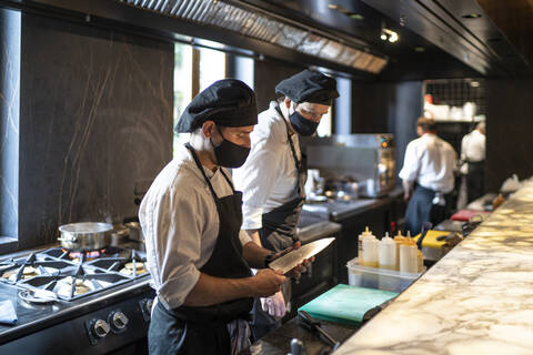 Köche mit Gesichtsschutzmasken arbeiten gemeinsam in einer Restaurantküche, lizenzfreies Stockfoto