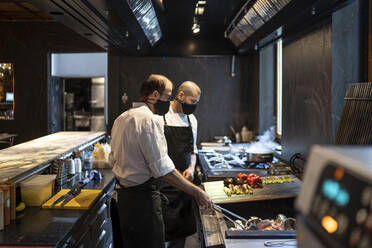 Chefs wearing protective face masks preparing grilled vegetables in restaurant kitchen - OCMF01502