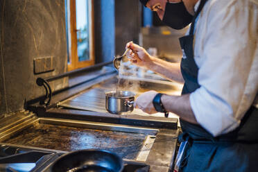 Koch mit Schutzmaske bei der Zubereitung einer Soße in einer Restaurantküche - OCMF01507