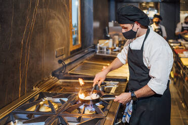 Chef wearing protective face mask preparing a dish in frying pan in restaurant kitchen - OCMF01511