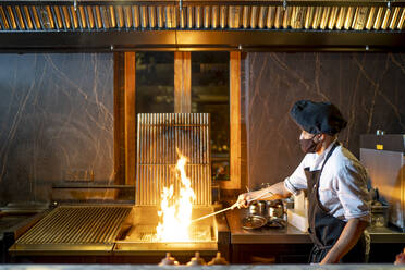 Chef wearing protective face mask preparing grill in restaurant kitchen - OCMF01520