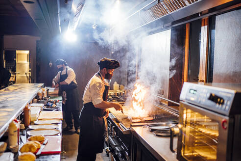 Köche mit Gesichtsschutzmasken bei der Zubereitung eines Gerichts in einer Restaurantküche - OCMF01521