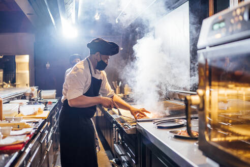 Koch mit Gesichtsschutzmaske bei der Zubereitung eines Gerichts in einer Restaurantküche - OCMF01523