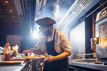 Koch mit Gesichtsschutzmaske bei der Zubereitung eines Gerichts in einer Restaurantküche - OCMF01524