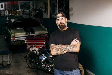 Tattooed mechanic standing with arms crossed in the garage and looking at camera. - ADSF03688