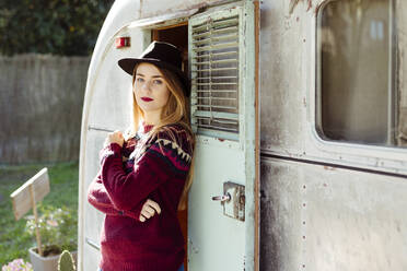 Schöne junge Frau in modischem Hut und Pullover hält die Arme verschränkt und schaut in die Kamera, während sie in der Nähe des Eingangs eines alten Wohnmobils steht - ADSF03694