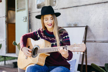 Beautiful young lady in stylish outfit sitting on chair near aged van and playing acoustic guitar - ADSF03698