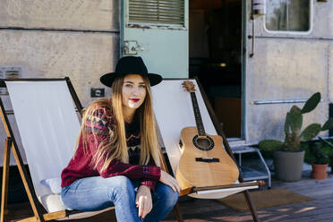 Beautiful young lady in stylish outfit sitting on chair near aged van and playing acoustic guitar - ADSF03699