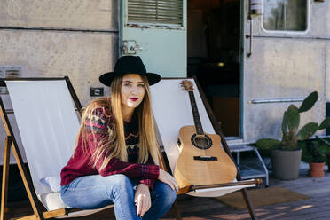 Schöne junge Frau in stilvollem Outfit sitzt auf einem Stuhl in der Nähe von alten van und spielt akustische Gitarre - ADSF03699