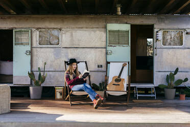 Charming young woman in stylish outfit reading interesting book while sitting on terrace near aged camper - ADSF03700